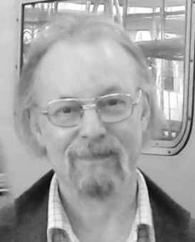 Black and white photo of a man with glasses and a beard, wearing a checked shirt and jacket, standing indoors.