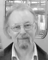Black and white photo of a man with glasses and a beard, wearing a checked shirt and jacket, standing indoors.