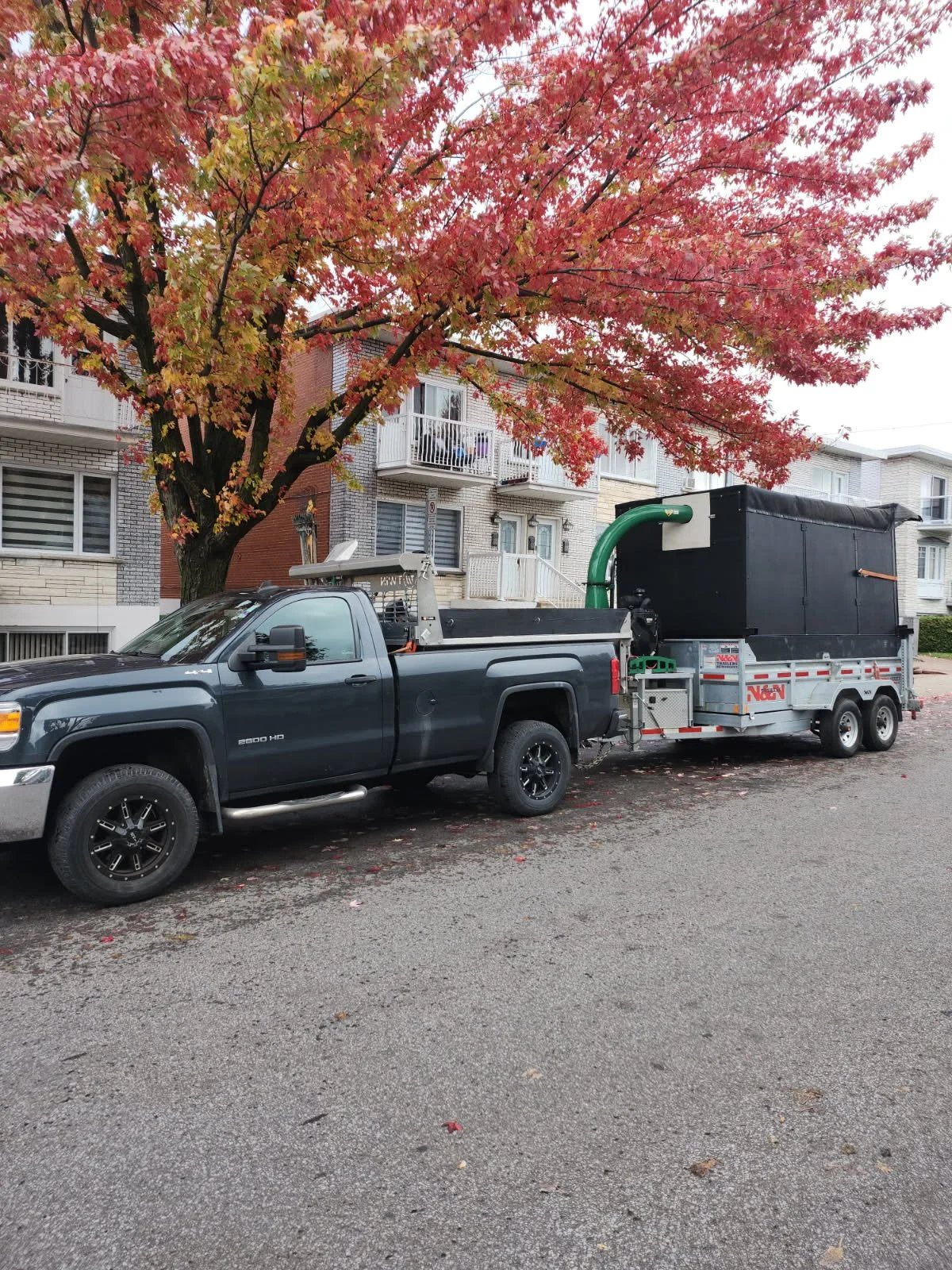 Black pickup truck with trailer parked on residential street, fall tree with red leaves, brick buildings in background.