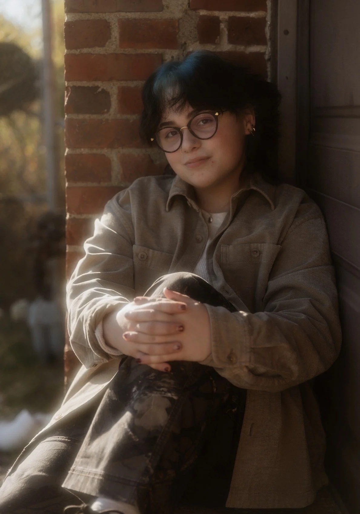 person with short black hair and glasses with tan jacket and jeans smiling at camera