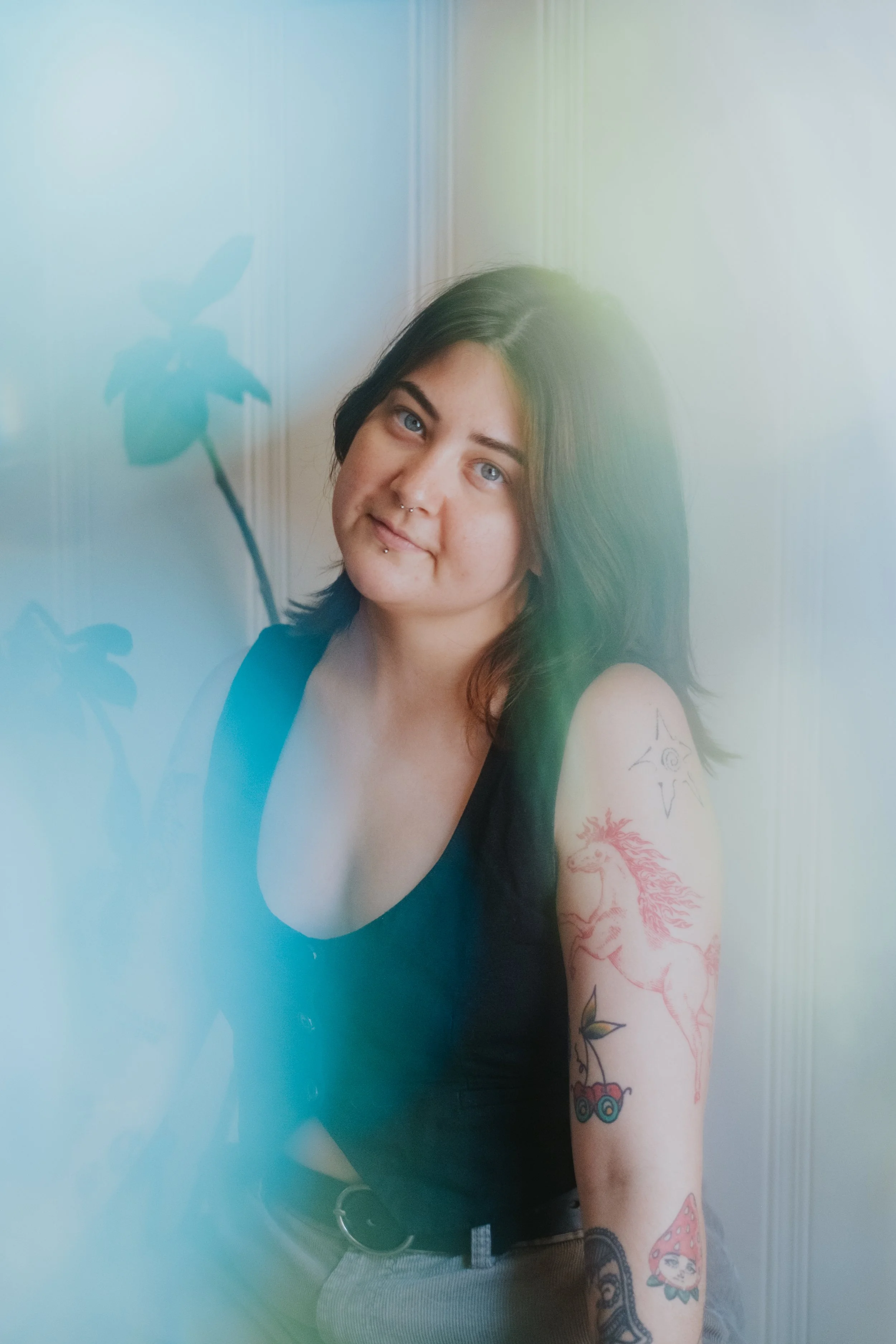 Young person with medium length dark hair and patchwork arm tattoos sitting in front of a white wall with plants