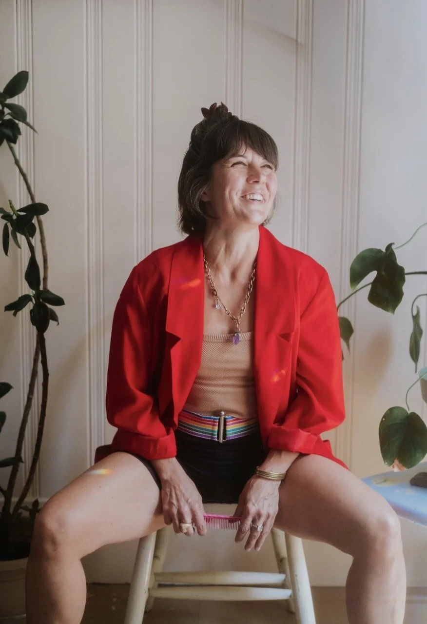 Women in red blazer and rainbow belt smiling seated near plants