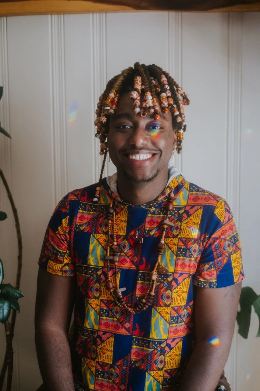 Person with beaded braided hair wearing patterned shirt, and large earrings, smiling against a plain background.