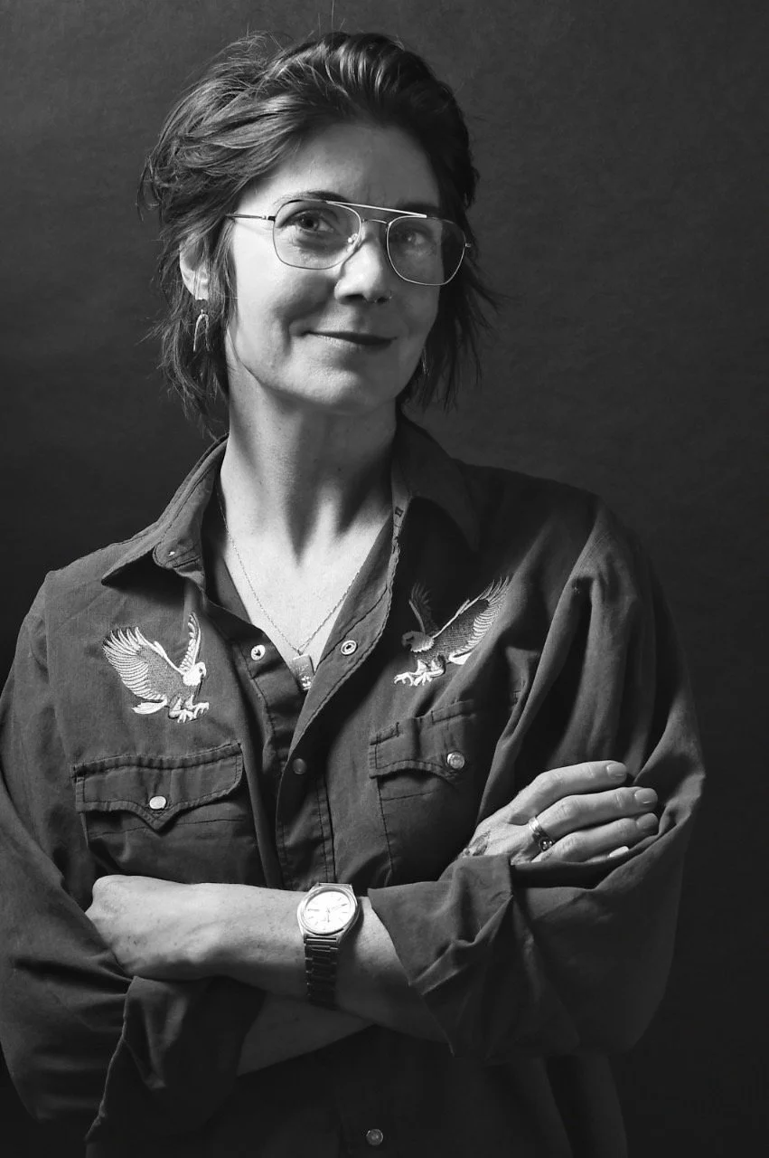 Person inside a hair salon smiling, wearing glasses, a red plaid shirt, with tattoos on arms and a red flower in hair. Salon has wooden shelves with products and a chandelier.