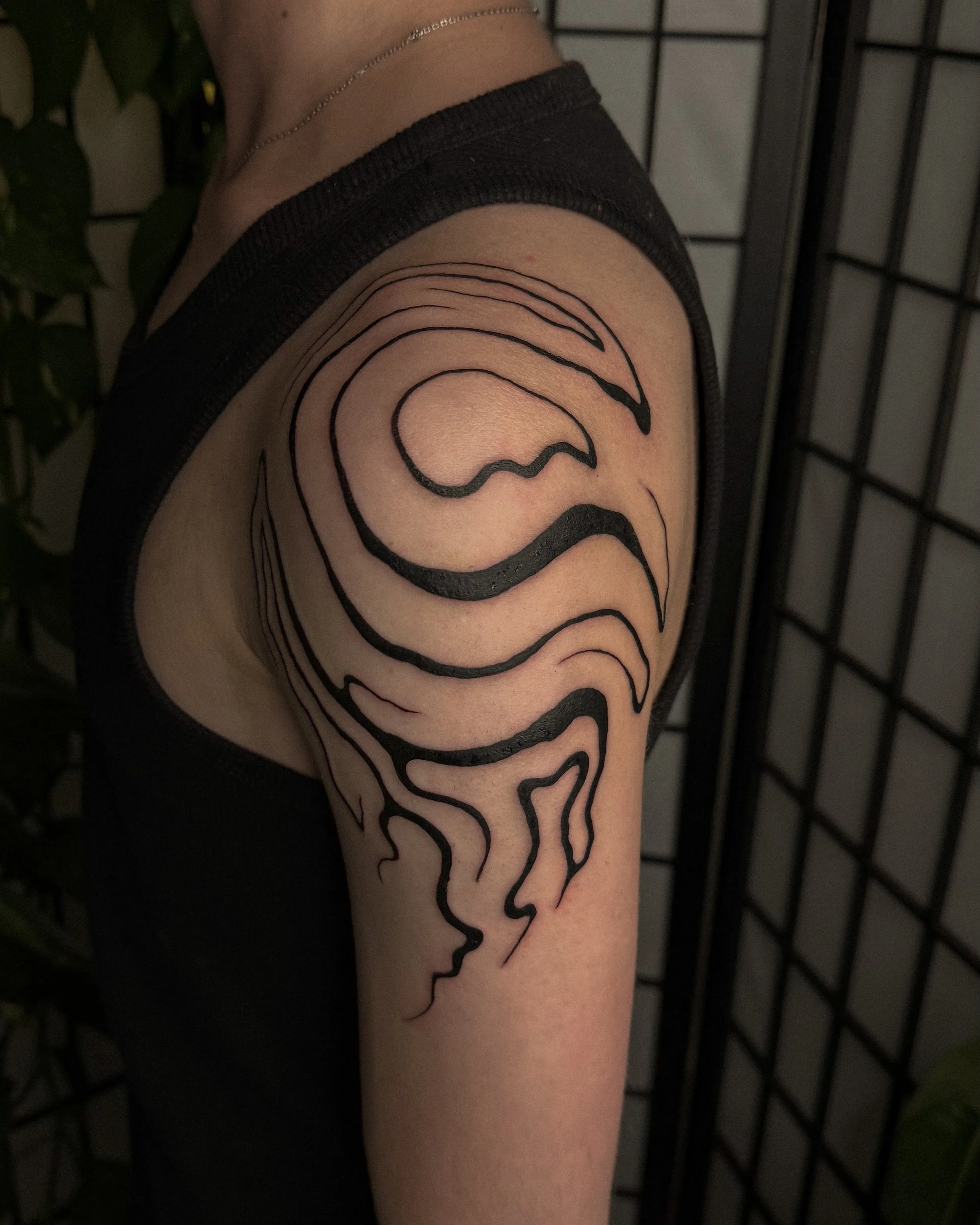 A person's upper arm with a black abstract line tattoo of a face outline. The individual is wearing a black tank top, a delicate chain necklace, and is standing against a background with a black metal grid and some green foliage.