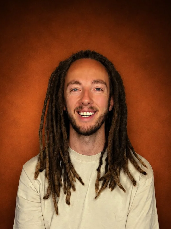 A man with long dreadlocks, a beard, and a big smile, wearing a cream-colored shirt against a brown background.