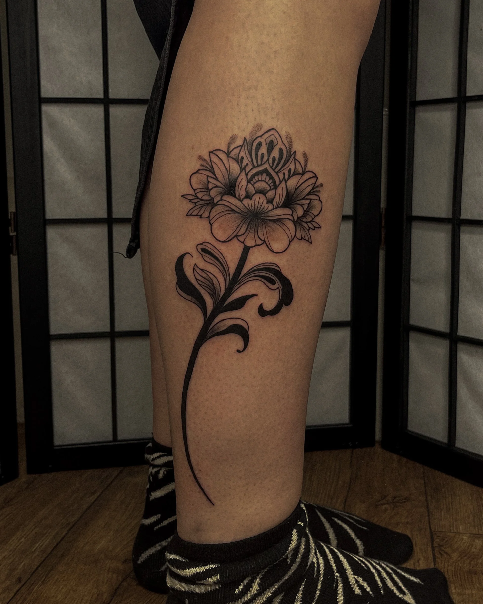 A person's arm with a detailed black and gray floral tattoo featuring a large flower with multiple petals and leaves, visible from the elbow to the wrist, with a background of black room dividers with lattice design.