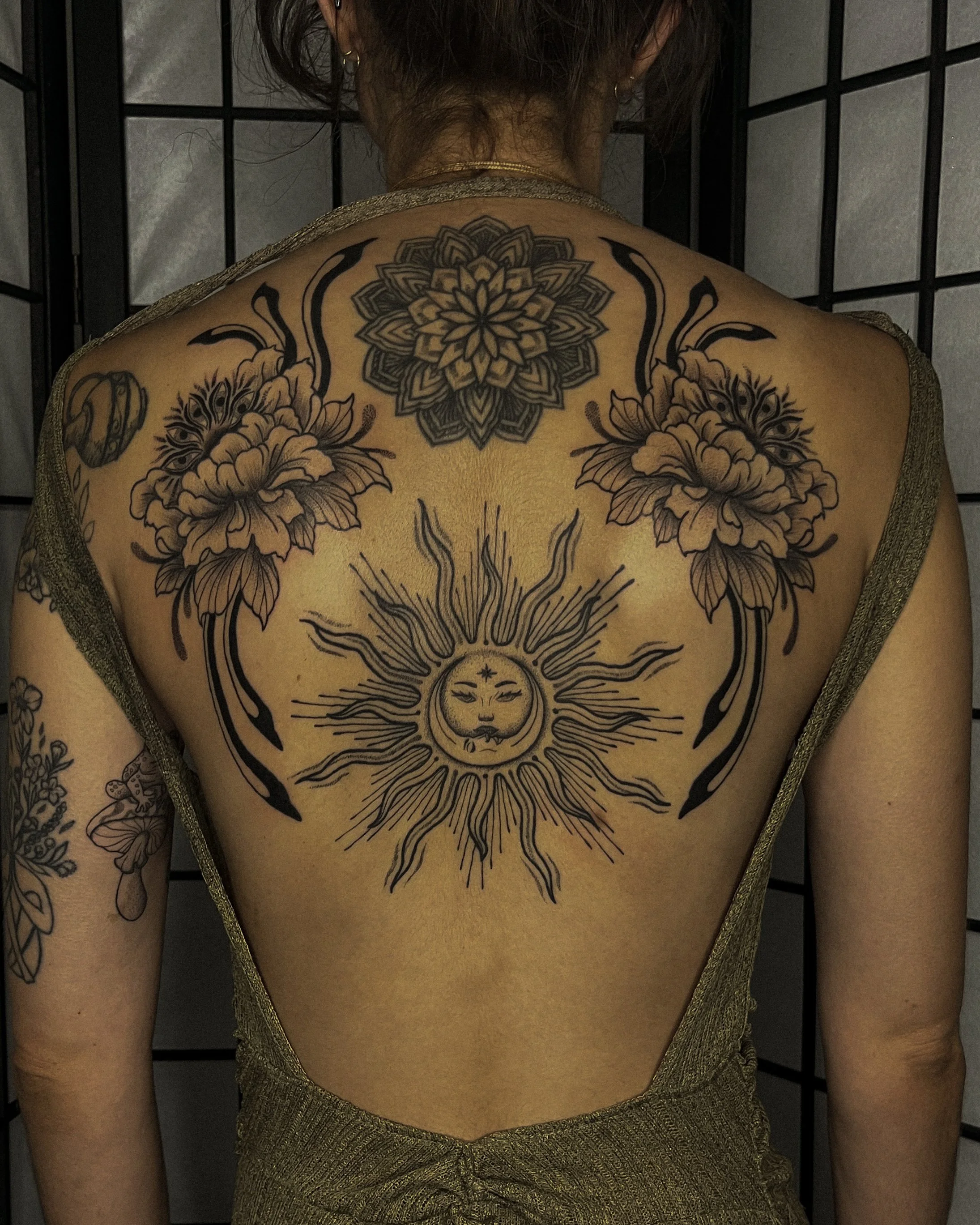 A person with tattoos on their back featuring a large mandala at the top, floral designs on the shoulders, and a sun and moon face in the center. The person is wearing a gold dress with thin straps and has earrings and necklaces. The background is a black grid-like pattern.