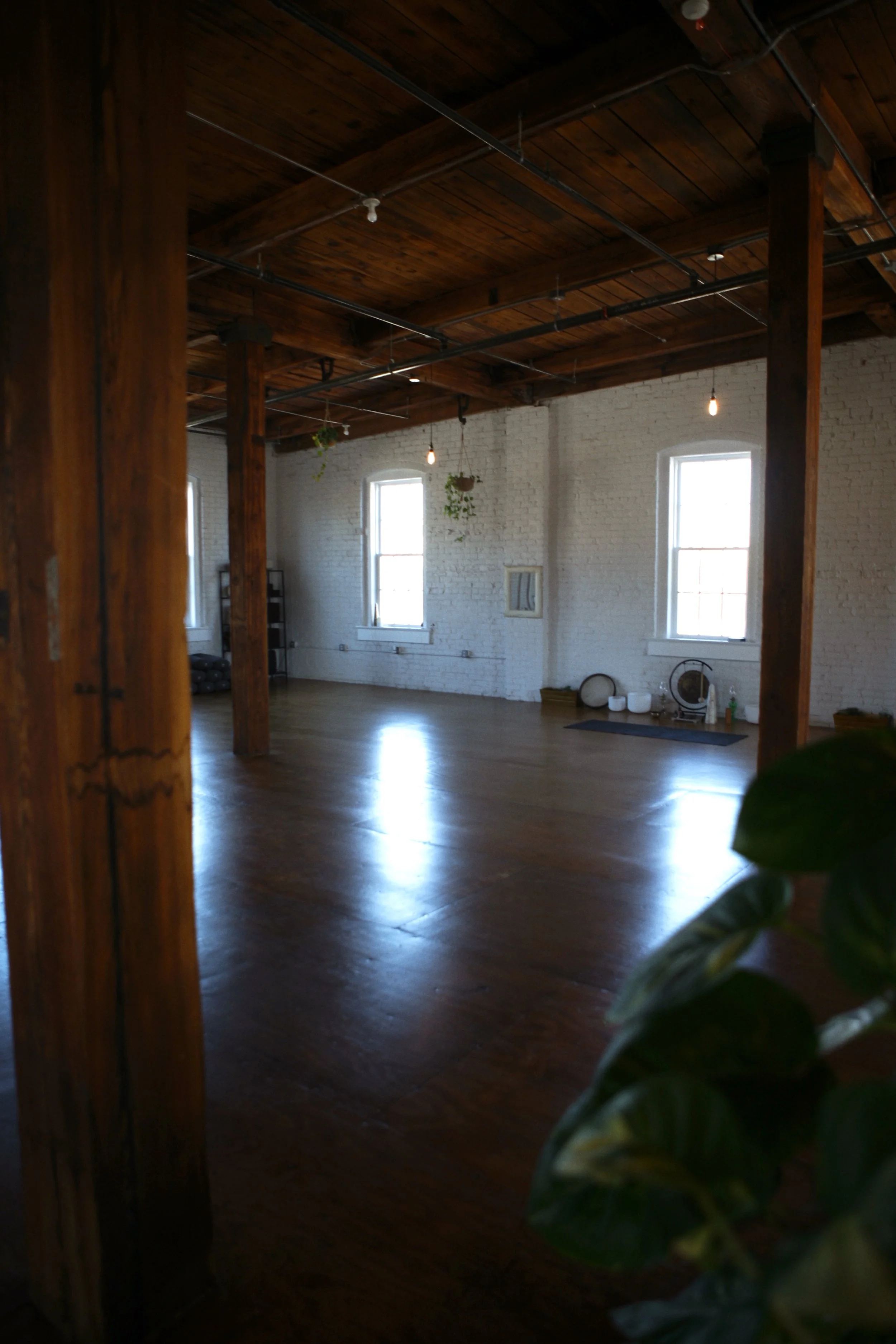 Empty industrial-style room with wooden beams, white brick walls, hardwood floors, two large windows, hanging light bulbs, potted plants, and some fitness equipment.