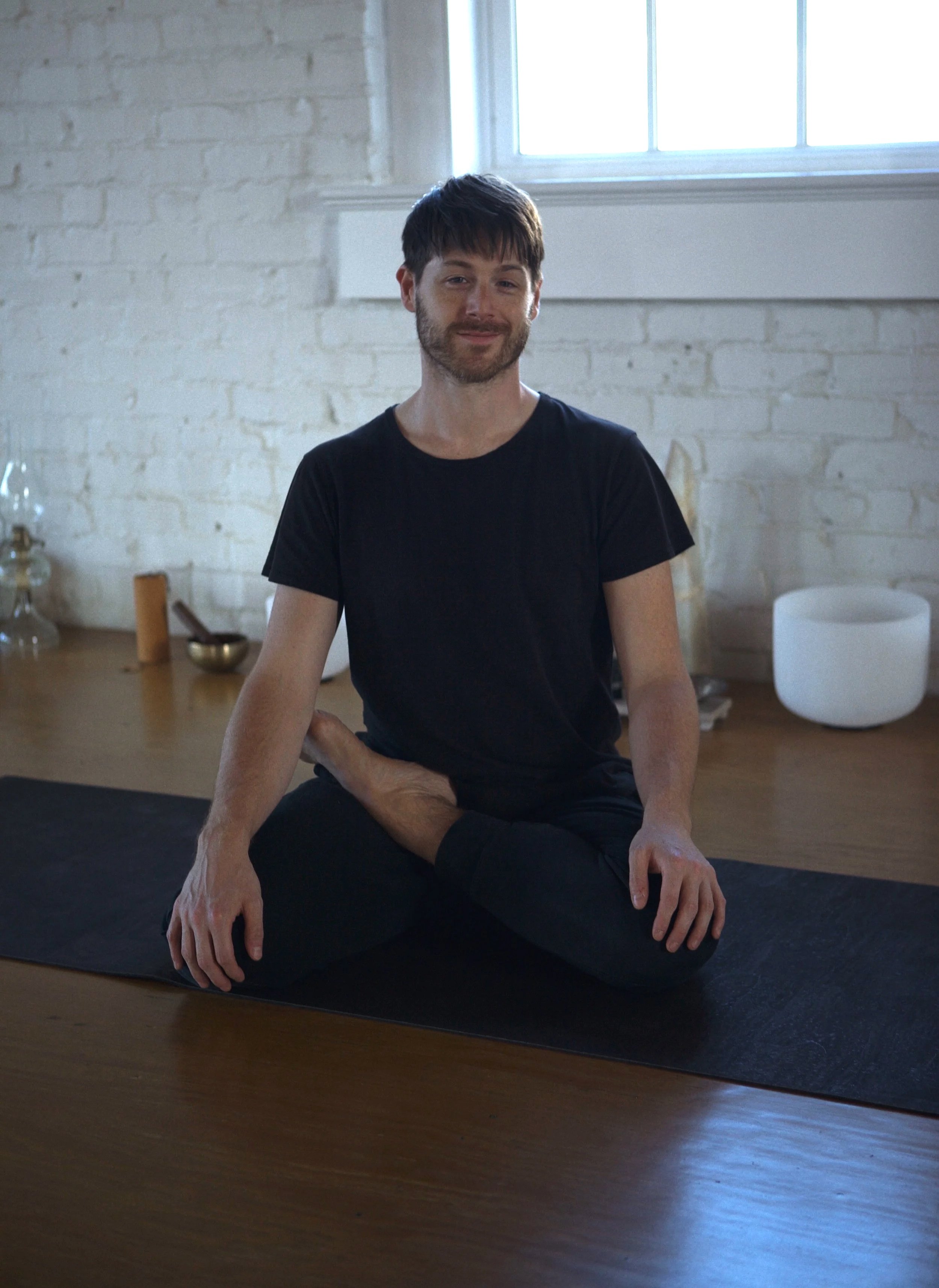A man practicing yoga or meditation on a black yoga mat in a bright room with a large window and white brick walls. He is sitting cross-legged and smiling.