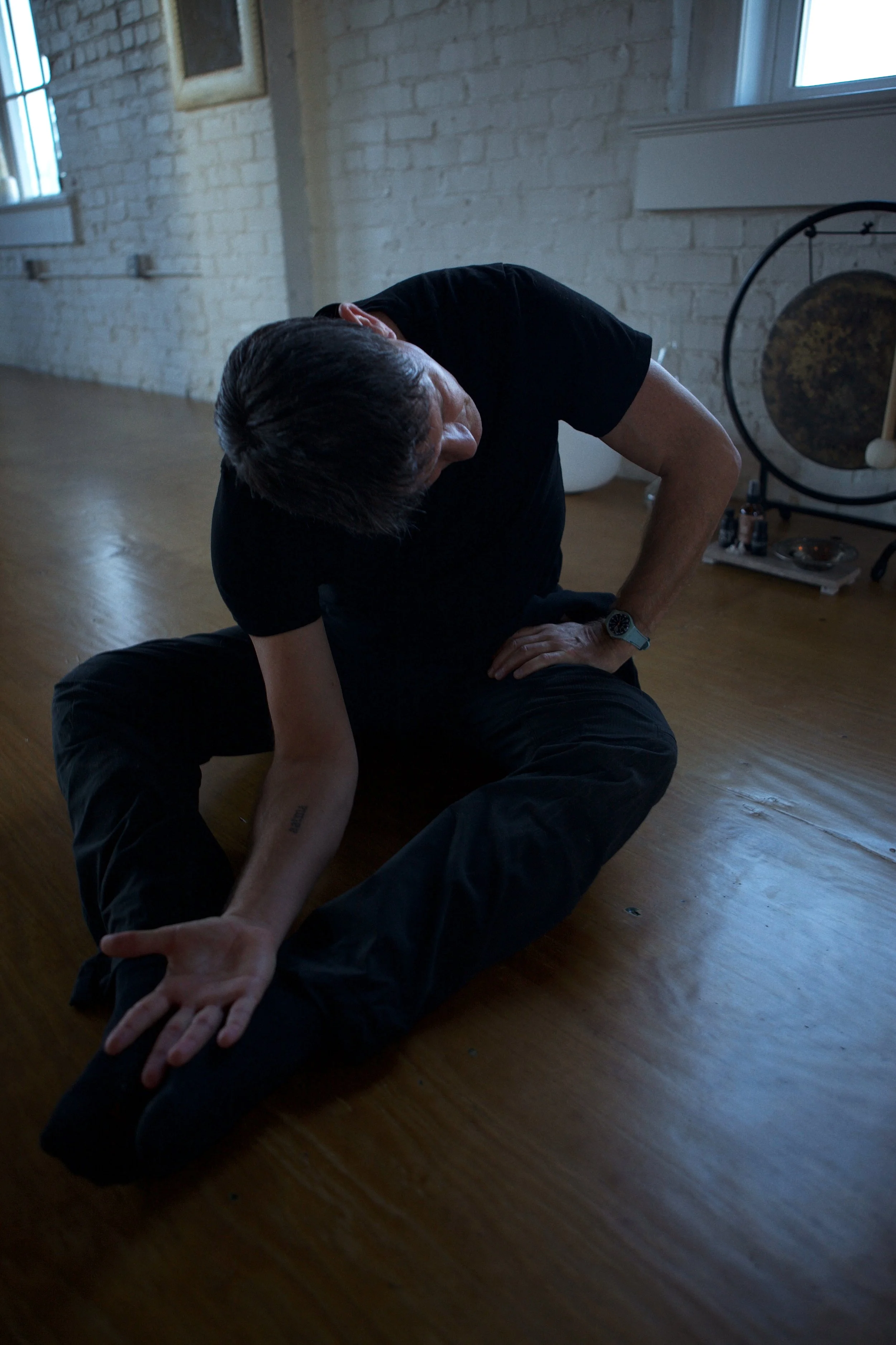A man practicing yoga or stretching on a wooden floor in a room with white brick walls, a window, and a gong in the background.