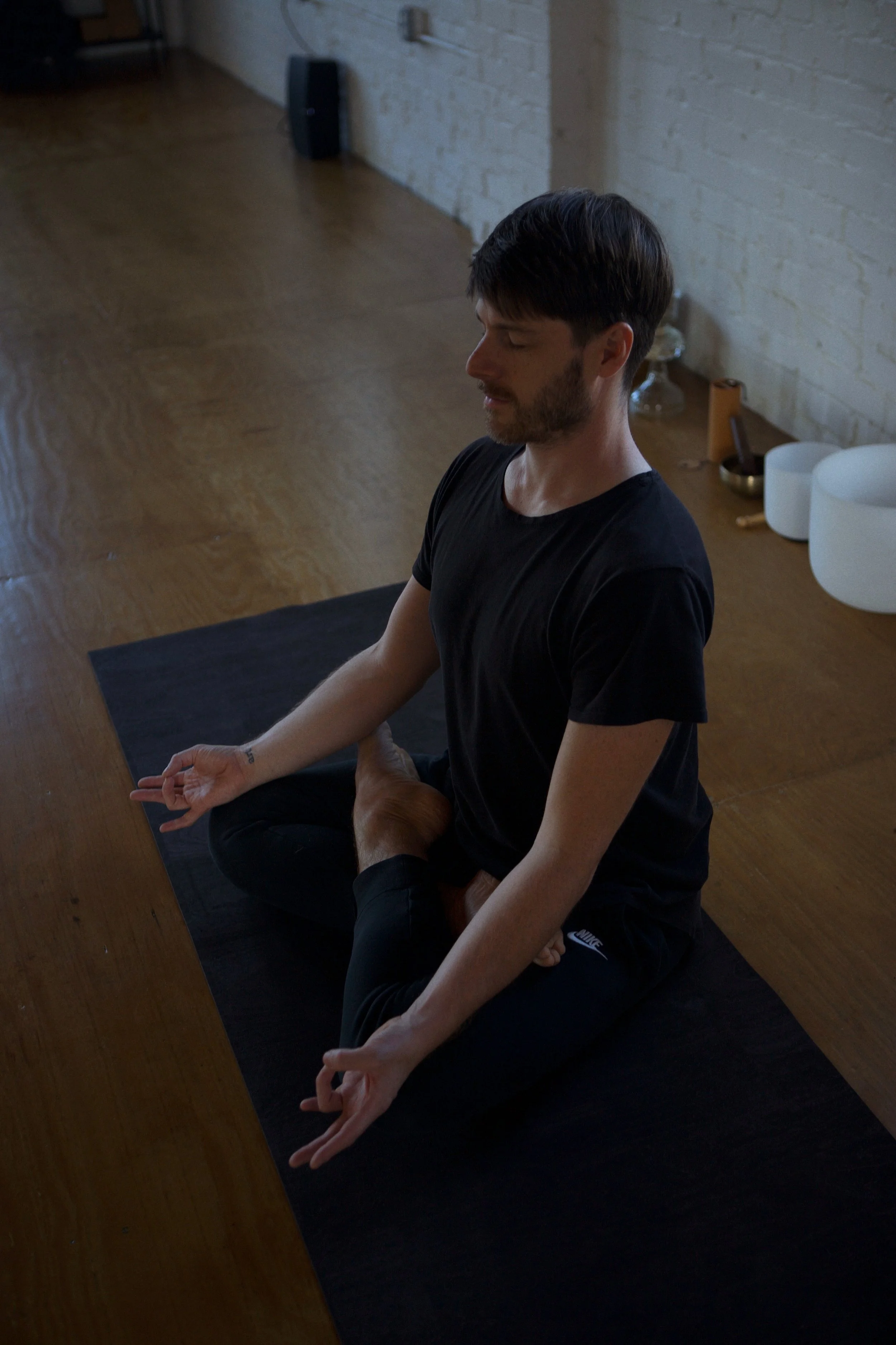 A man practicing yoga in a meditation pose, sitting cross-legged on a black mat in a room with wooden flooring and a brick wall.
