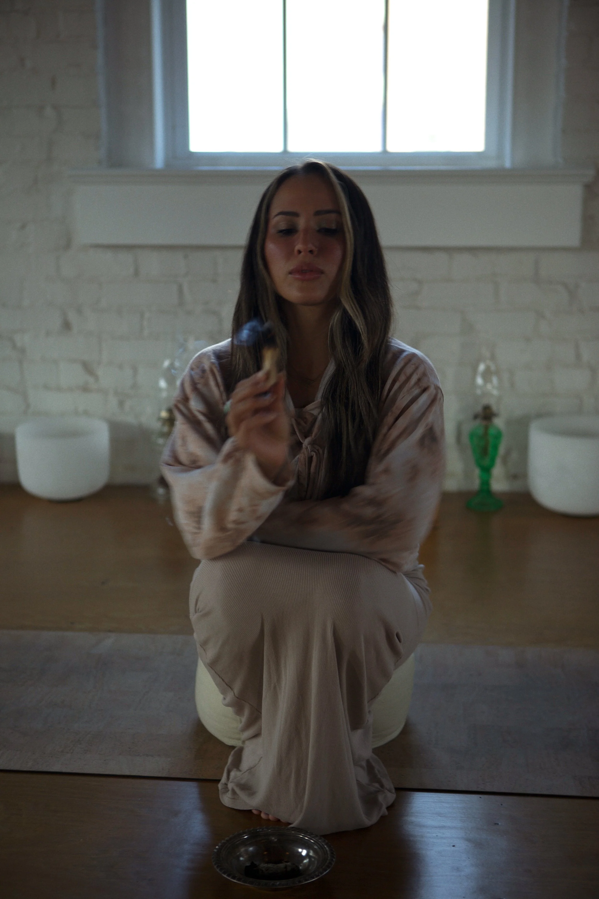 A woman with long, wavy hair sitting cross-legged on a cushion, holding an object in her hand, in a room with white brick walls, a window overhead, and various decorative items in the background.