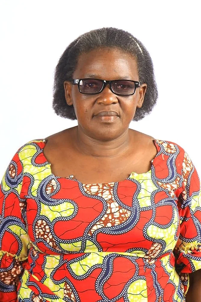An African woman wearing dark sunglasses and a colorful, patterned dress with red, yellow, and blue designs, standing against a plain white background.