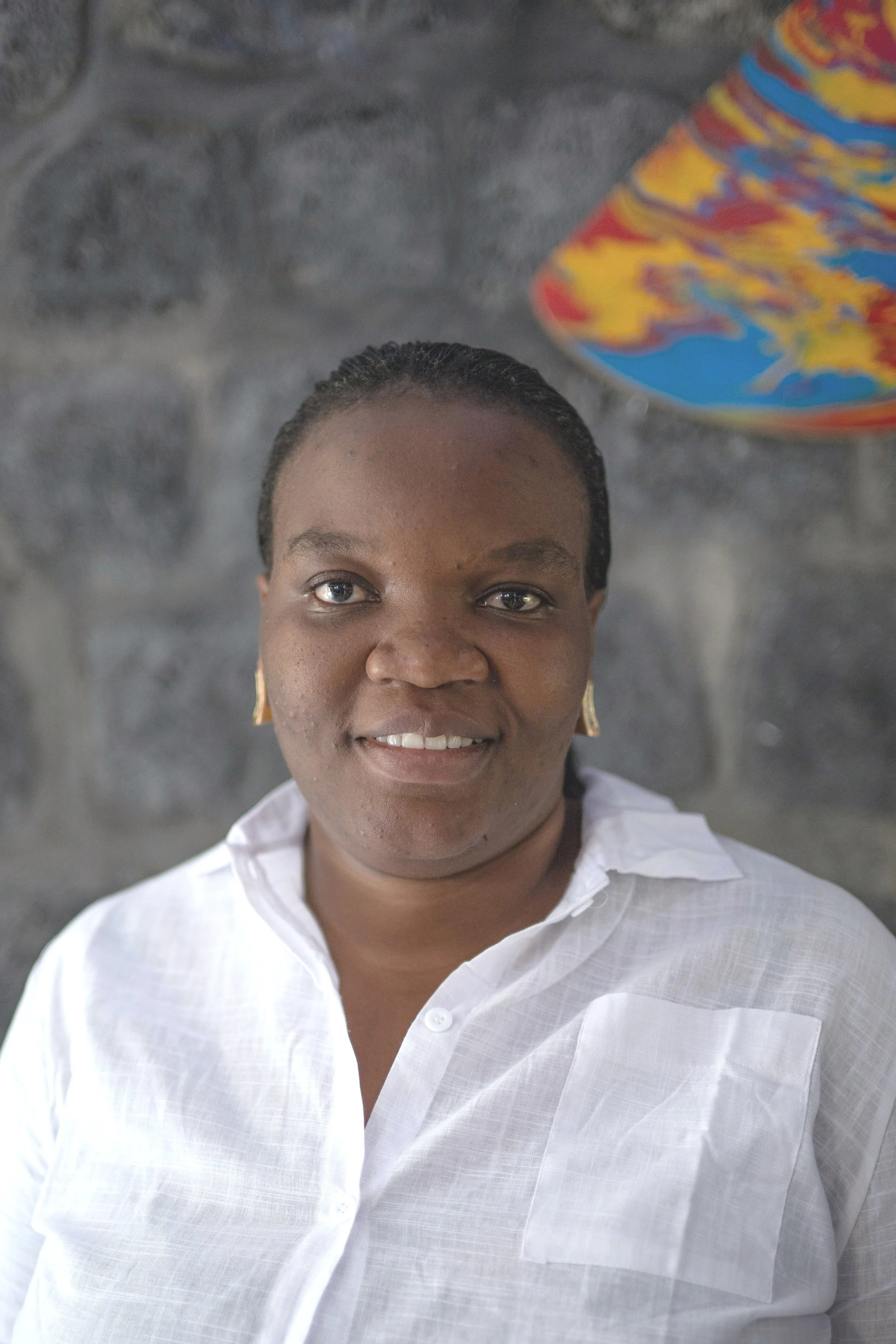 A woman wearing a white shirt and gold earrings, smiling at the camera, with a stone wall and a colorful object in the background.