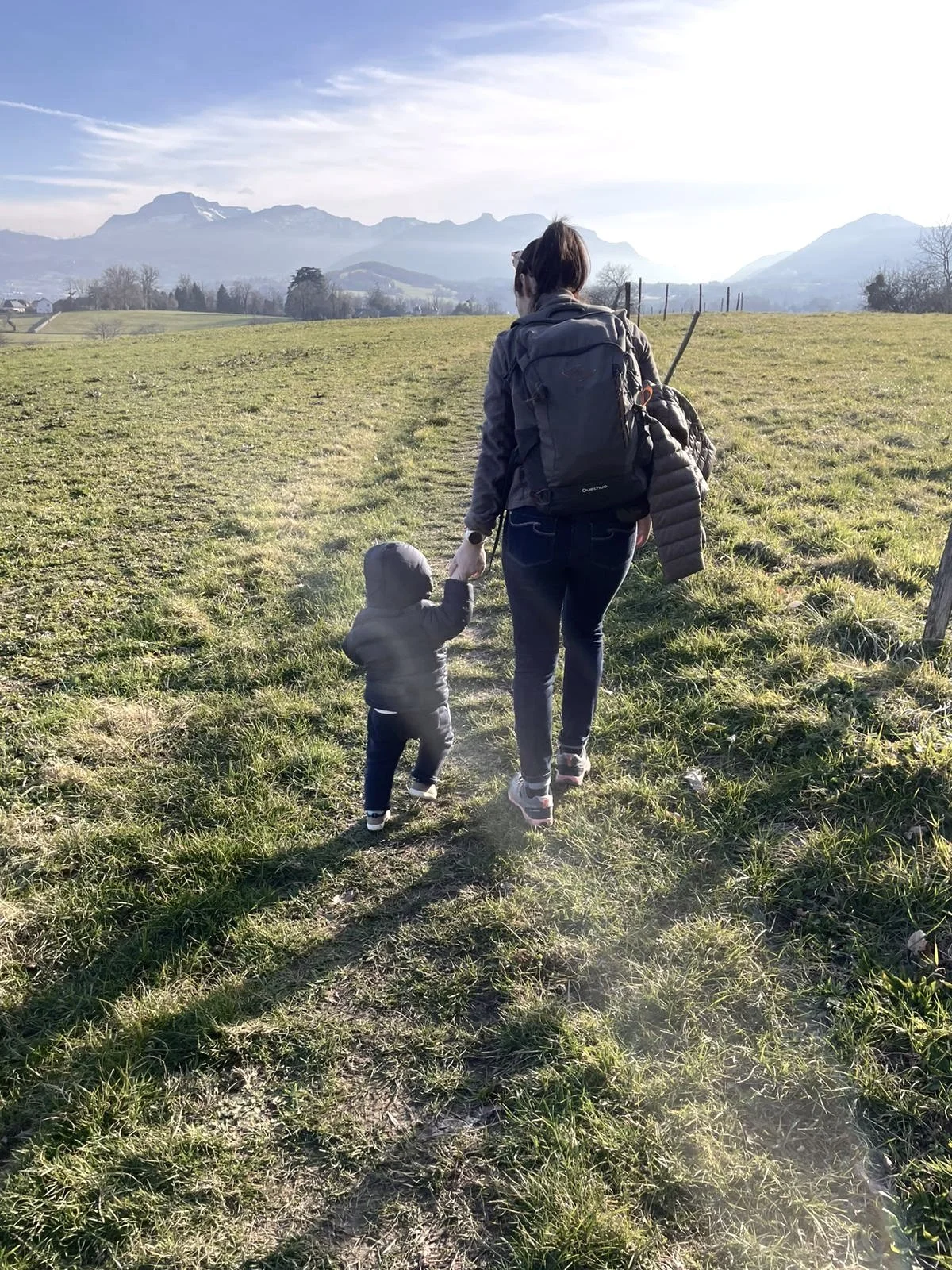Une personne adulte et un enfant marchent main dans la main sur un chemin de campagne, avec des montagnes en arrière-plan.