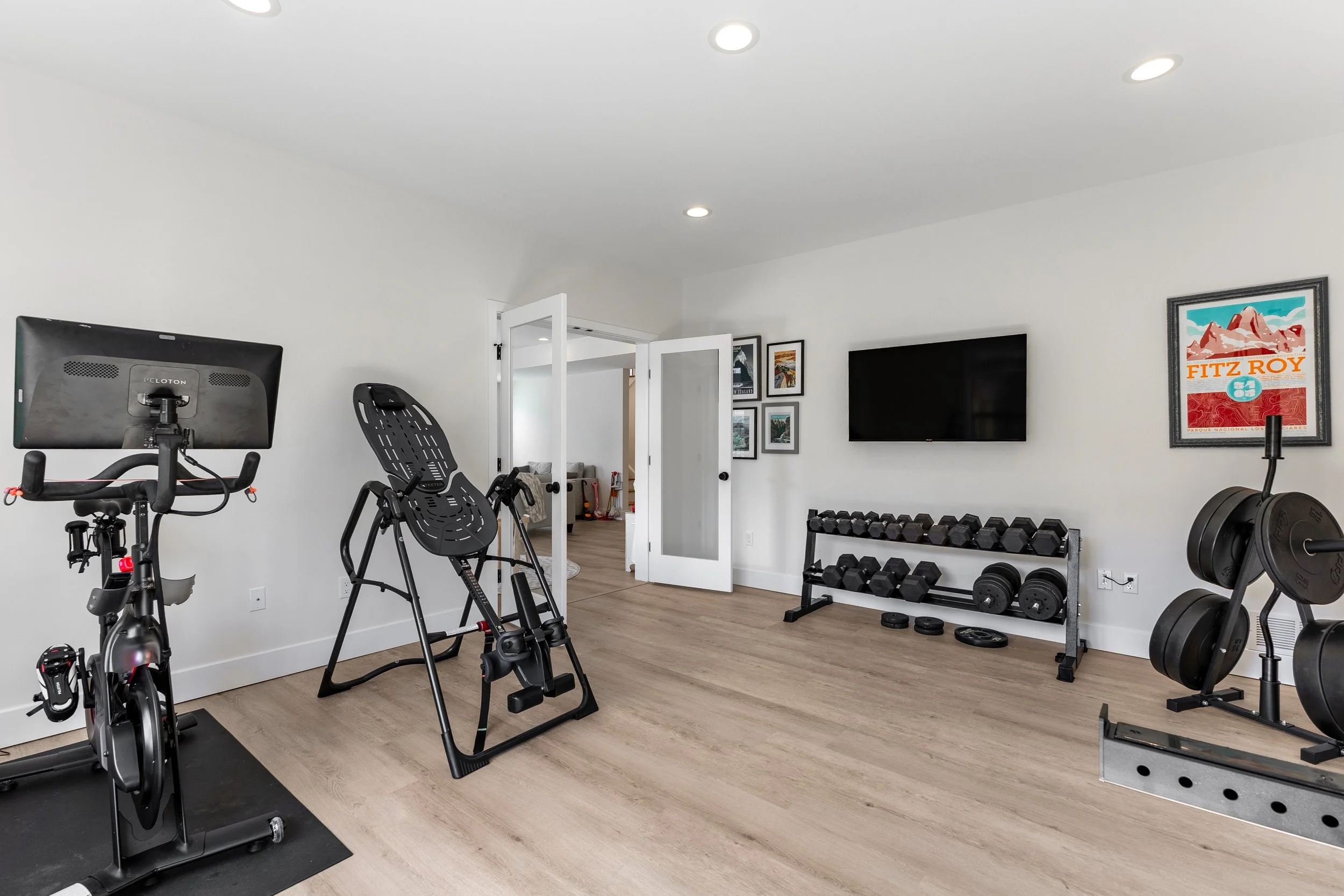 Home gym with exercise bike, rowing machine, dumbbells, weight rack, and a flat screen TV on white walls.