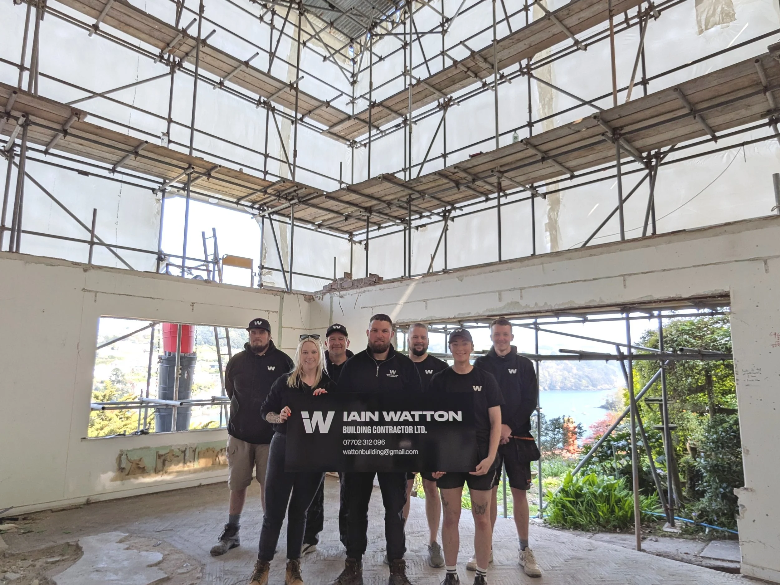 Group of six people in construction site holding a sign, surrounded by scaffolding. The sign reads "Iain Watton Building Contractor Ltd.". The site has unfinished walls and a view of trees and water outside.