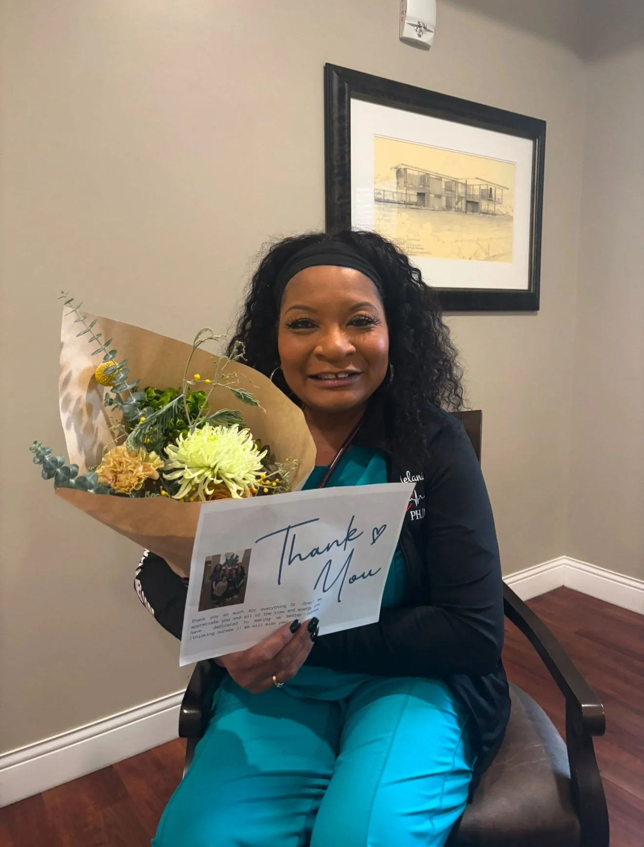 A woman with curly black hair, wearing a black jacket and teal scrubs, is smiling and holding a bouquet of flowers and a thank you card. She is sitting in a room with beige walls, a framed picture of a building on the wall, and wood flooring.