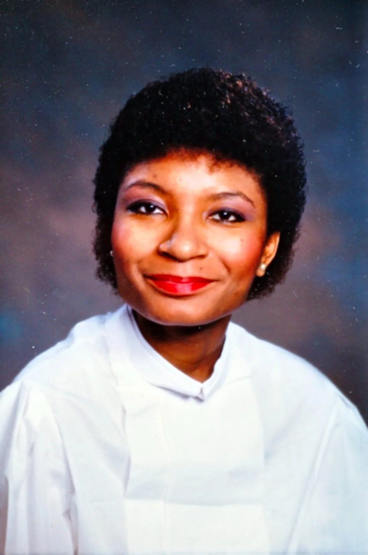 Studio portrait of an African American woman with short curly hair, wearing a white top, bright red lipstick, and earrings, against a dark, subtly speckled background.