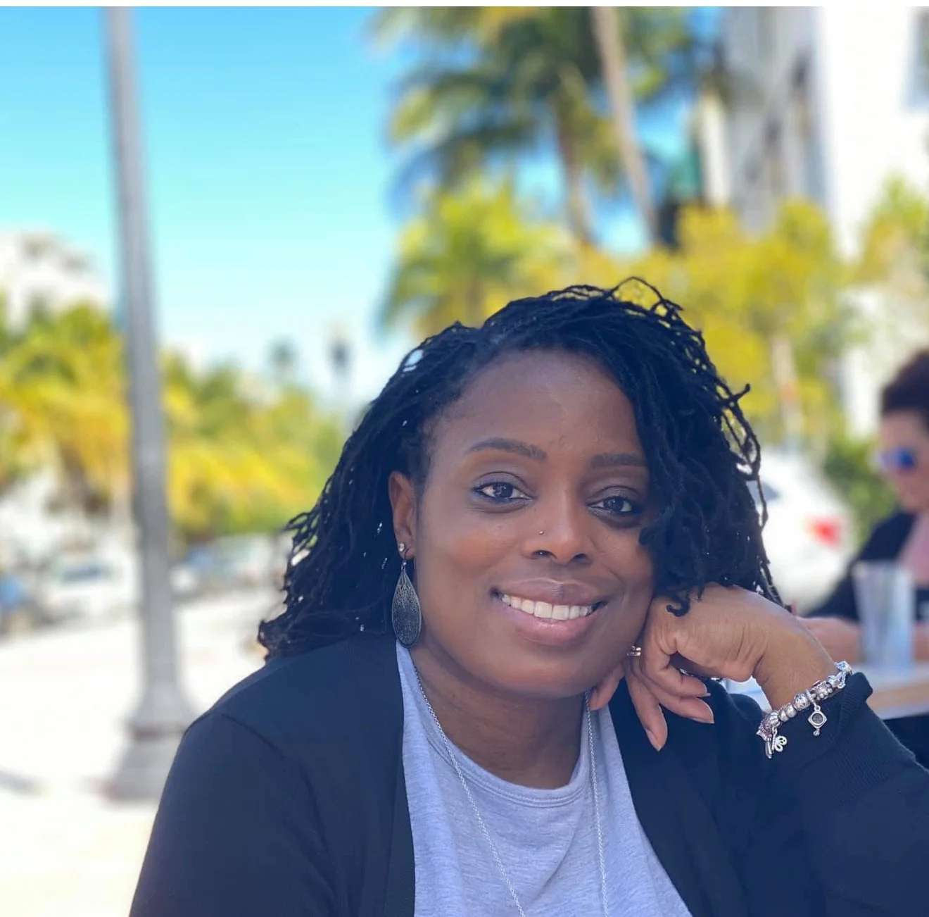A woman with dark, curly dreadlocks smiling, resting her chin on her hand outdoors with palm trees and a person in the background.