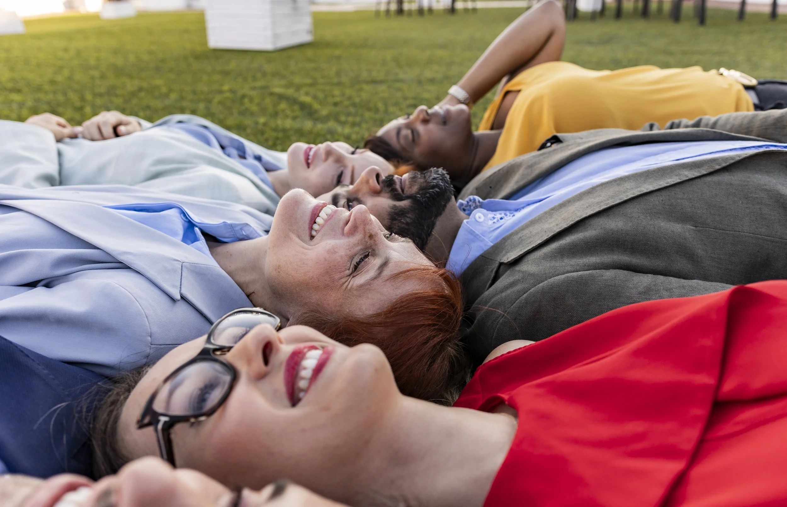 Group of five diverse adults lying on the grass outdoors, smiling, and relaxing together.