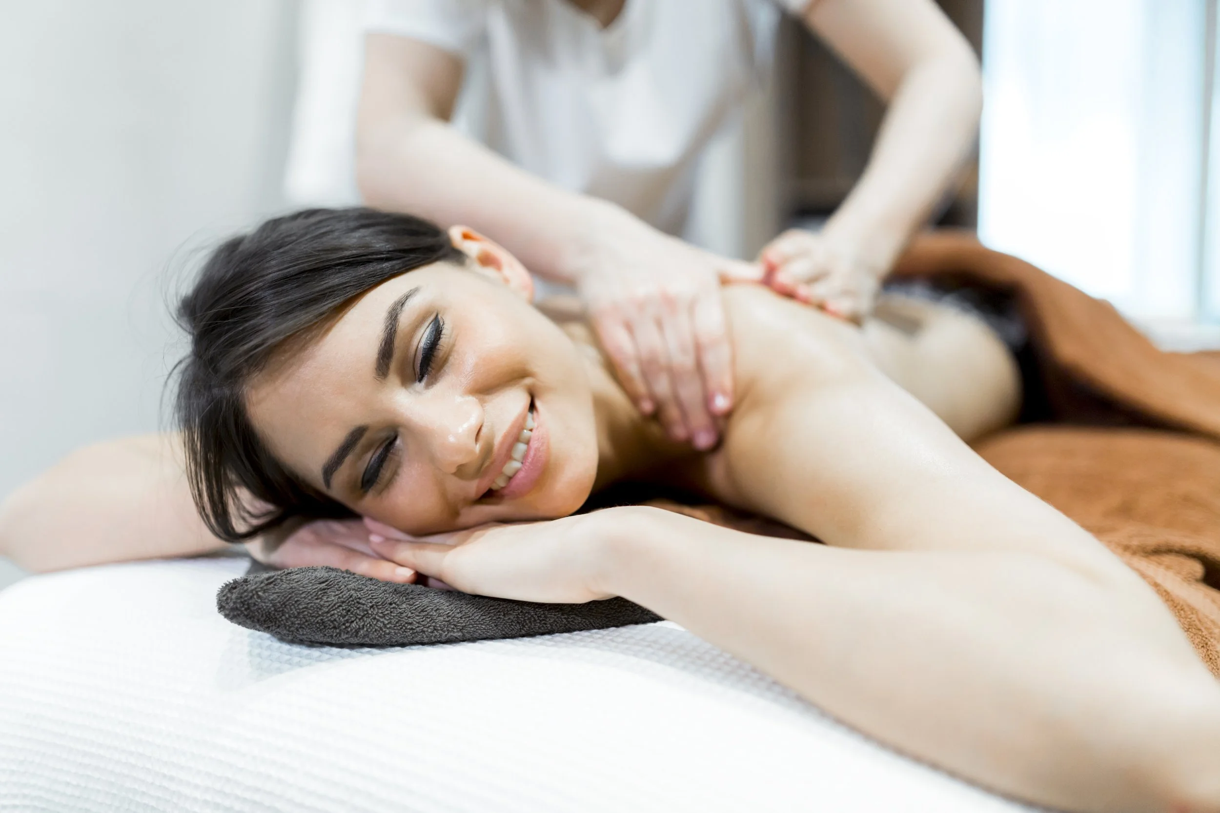A woman receiving a massage and smiling, lying face down on a massage table with her head turned to the side, eyes closed, and resting on her arm.