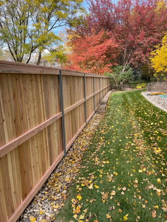 A new wooden privacy fence along a green grass lawn with fallen autumn leaves, trees with colorful fall foliage in the background, and a gravel border. Great Plains Fence is an expert fence installation and repair company in Sioux Falls, South Dakota