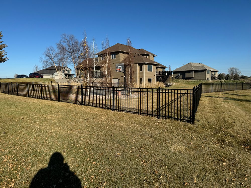 A backyard with a black metal fence, some leafless trees, and a large house with beige siding and multiple levels, under a clear blue sky. Great Plains Fence is an expert fence installation and repair company in Sioux Falls, South Dakota.