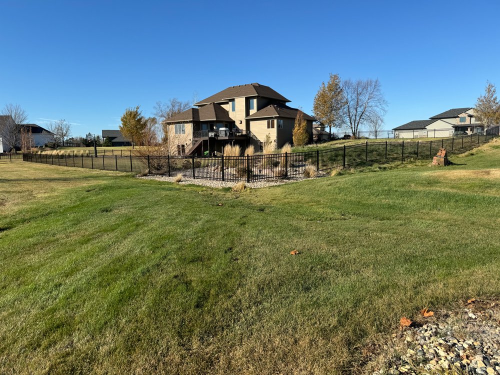 A suburban yard with green grass, a black fence, and a large beige house with a deck. Great Plains Fence is an expert fence installation and repair company in Sioux Falls, South Dakota.