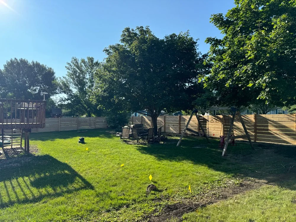 Backyard with green grass, several trees providing shade, patio furniture, a swing set, and yellow markers placed in the ground. Great Plains Fence is an expert fence installation and repair company in Sioux Falls, South Dakota.