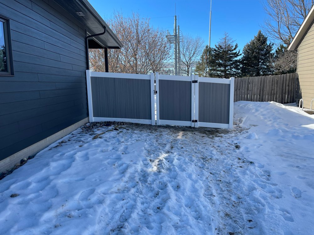 View of a backyard with snow-covered ground, a blue house on the left, a wooden fence in the background, and a gray gate with white trim in the center. Great Plains Fence is an expert fence installation and repair company in Sioux Falls, South Dakota