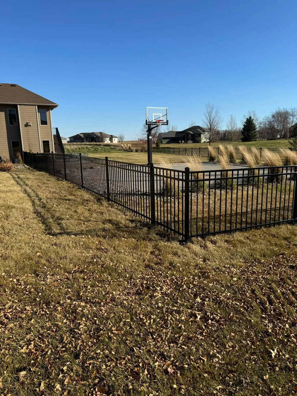 A fenced basketball court with a hoop, located next to a house with brown siding and a lawn. Great Plains Fence is an expert fence installation and repair company in Sioux Falls, South Dakota.