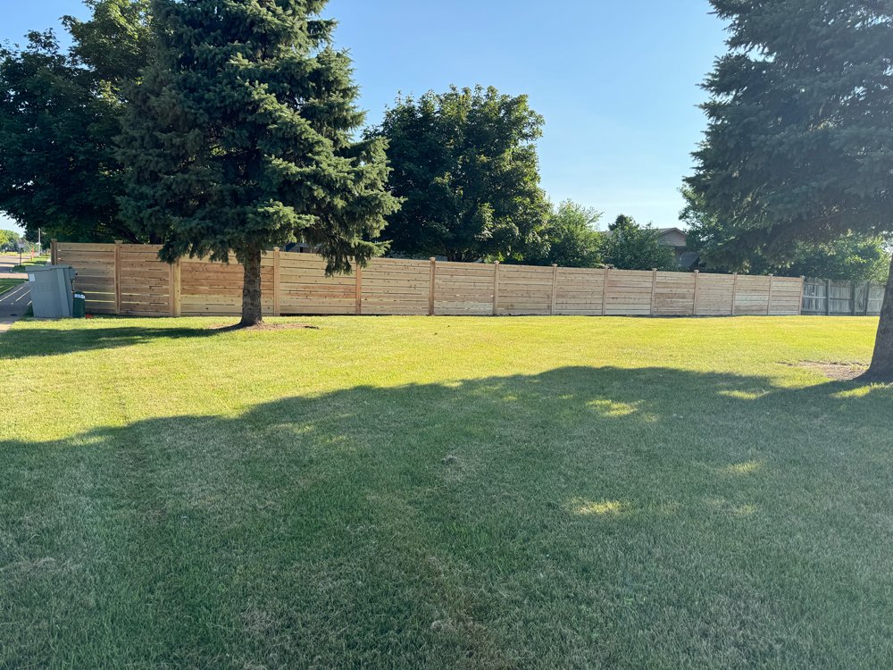 A grassy backyard with a wooden fence, large trees, and a blue sky. Great Plains Fence is an expert fence installation and repair company in Sioux Falls, South Dakota.