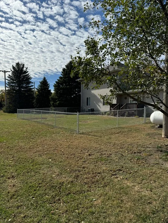 A residential backyard with a white house, a chain-link fence, green grass, and trees under a partly cloudy sky. Great Plains Fence is an expert fence installation and repair company in Sioux Falls, South Dakota.