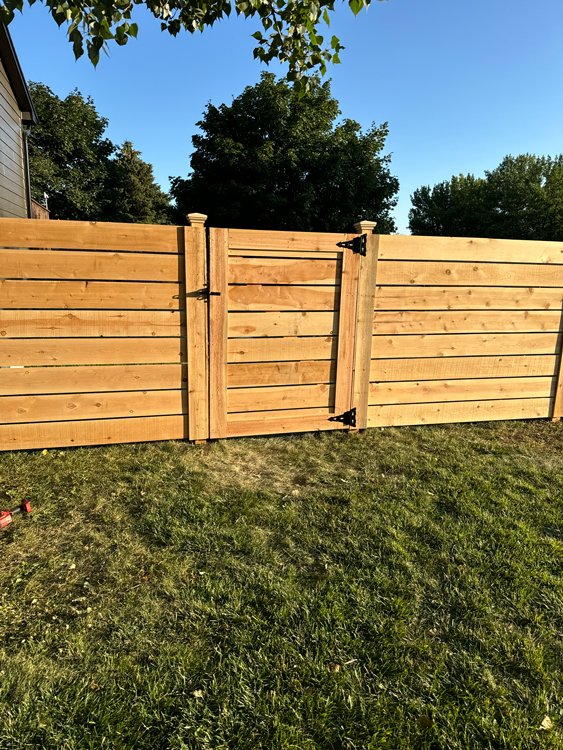 A wooden fence with a gate, situated on a grassy lawn with trees in the background under a clear blue sky. Great Plains Fence is an expert fence installation and repair company in Sioux Falls, South Dakota.
