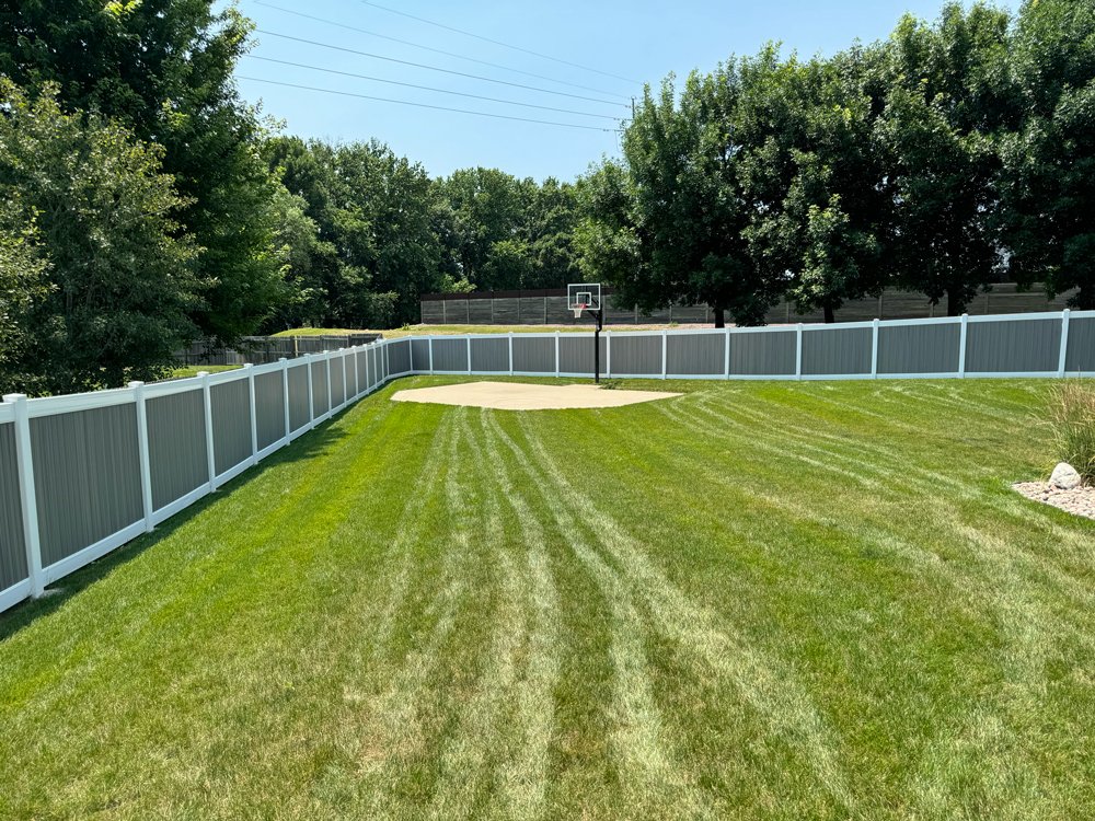 A backyard with a well-maintained lawn, enclosed by a white fence, featuring a basketball hoop and a sandy court area, surrounded by trees and a clear blue sky. Great Plains Fence is an expert fence installation and repair company in Sioux Falls, SD.