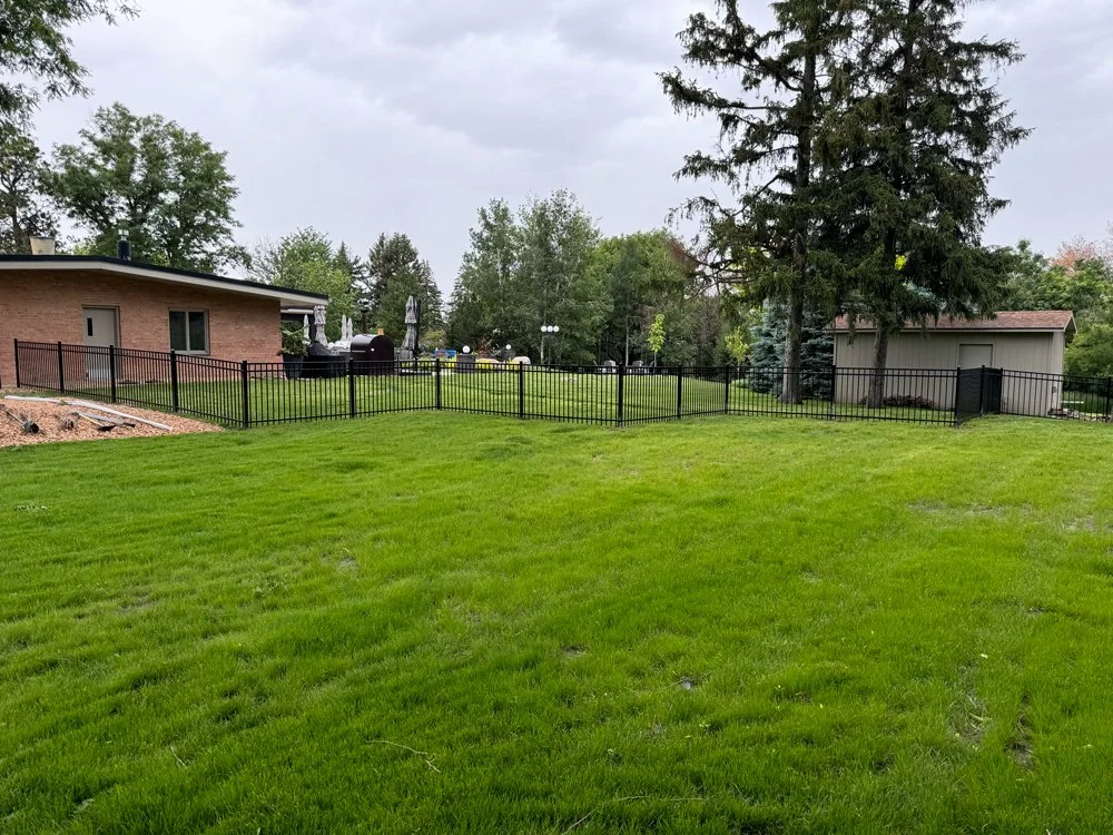 A backyard with a large green lawn, surrounded by a black metal fence. There are a brick house and a gray shed, with trees and cloudy sky in the background. Great Plains Fence is an expert fence installation and repair company in Sioux Falls, SD.