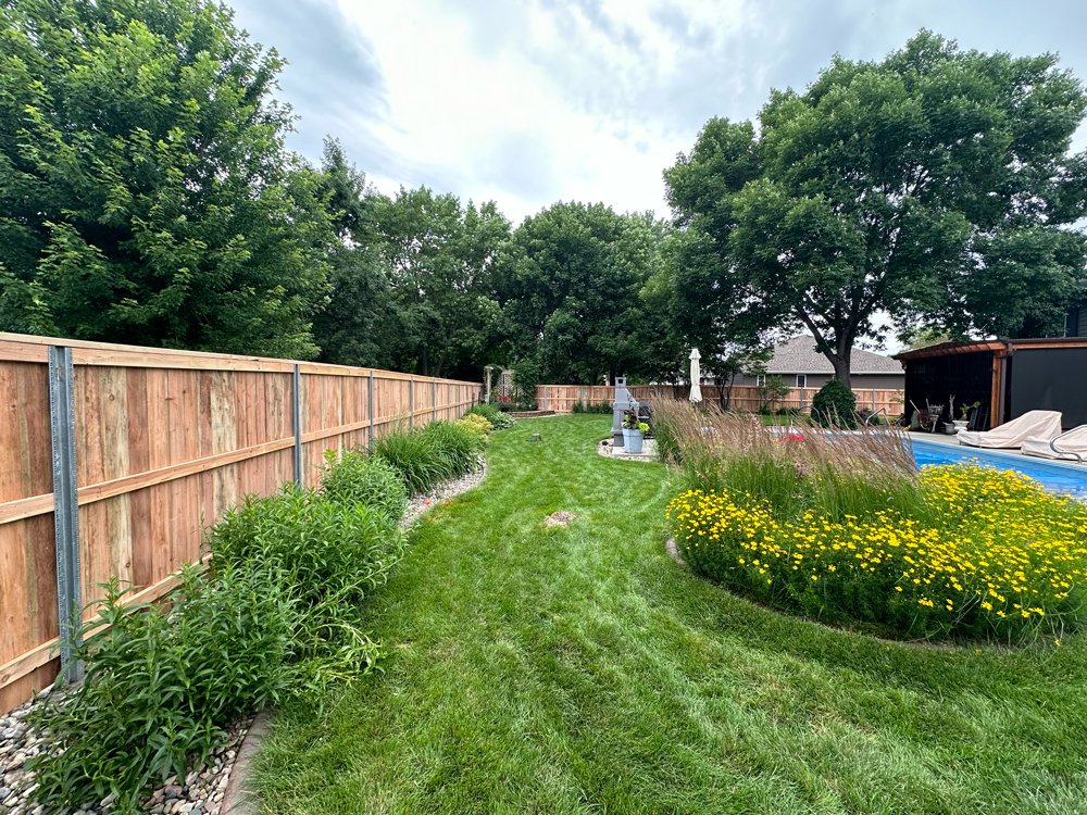 A backyard with a wooden fence on the left side, well-maintained green grass, and a flower bed on the right with yellow flowers and ornamental grasses. Great Plains Fence is an expert fence installation and repair company in Sioux Falls, South Dakota