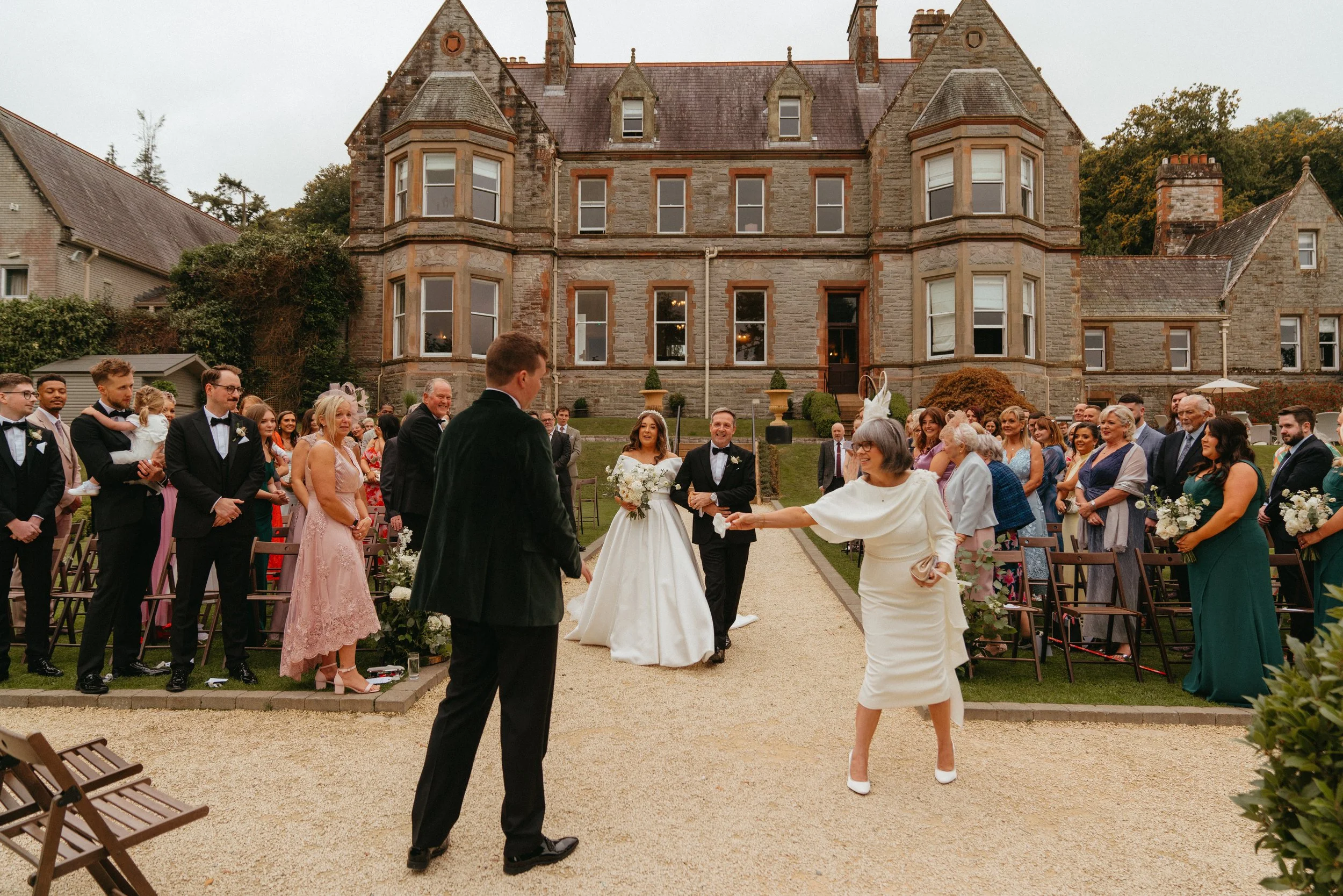 Outdoor wedding ceremony, bride walking down the aisle with guests seated on the sides, historic building in the background.