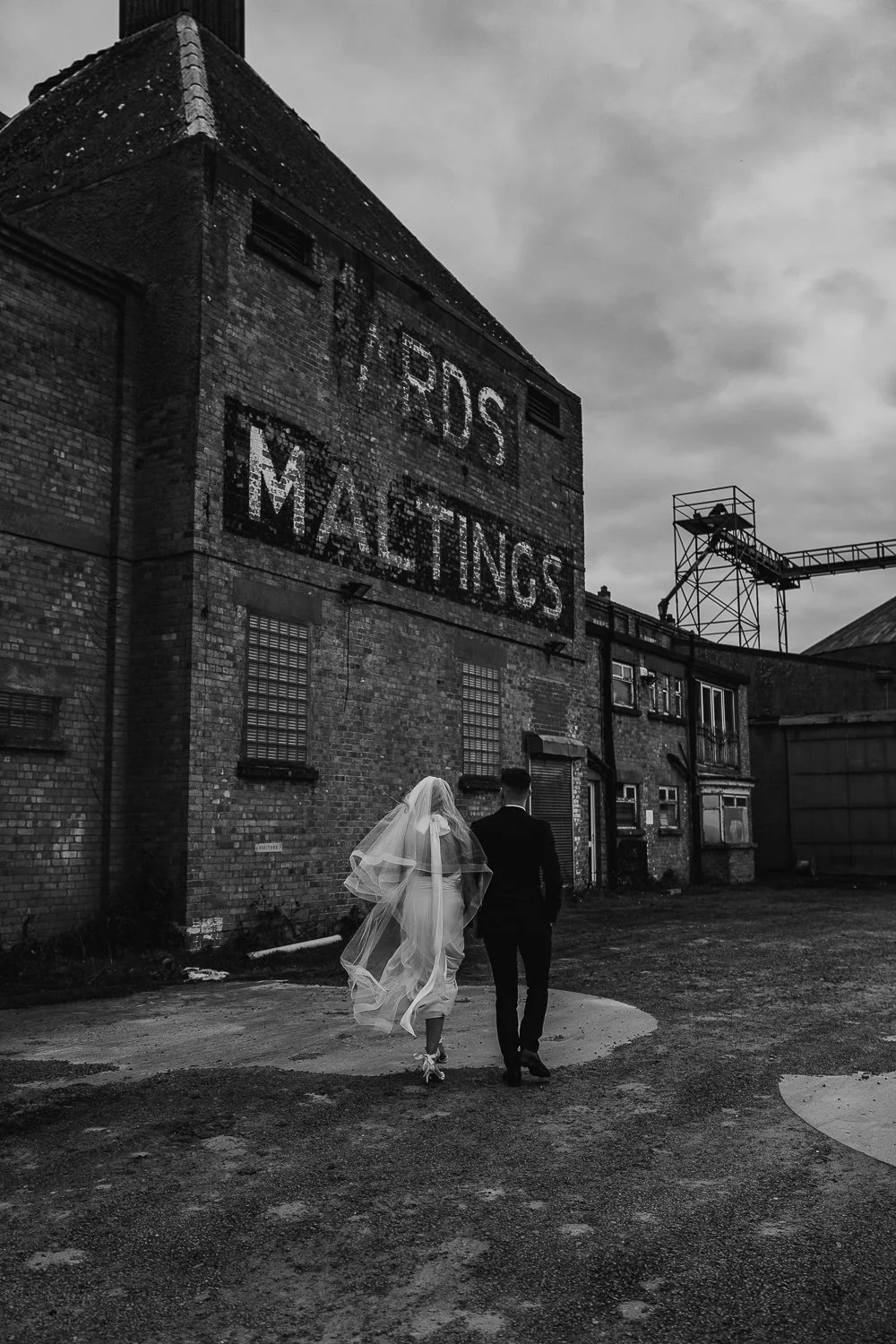 Black and white photo of a couple walking by an industrial building with "MALTINGS" sign.