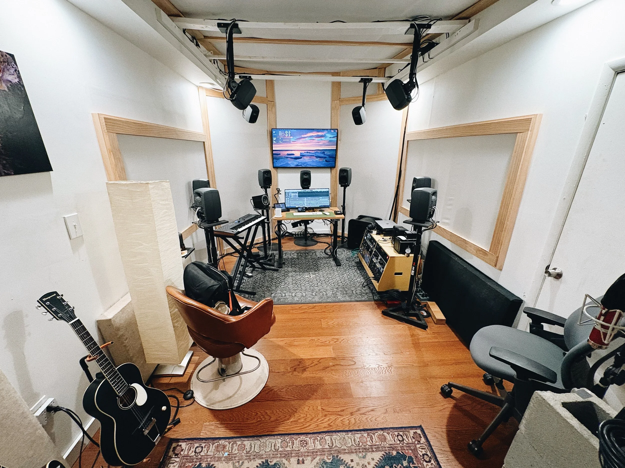 Recording studio with various audio equipment, a TV monitor, guitars, chairs, and soundproofing panels.