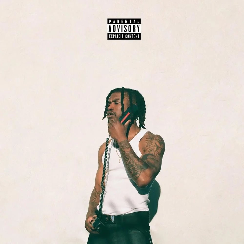 Young man with tattoos holding a phone to his ear, wearing a white tank top and black pants, standing against a plain white wall, with a parental advisory label at the top.