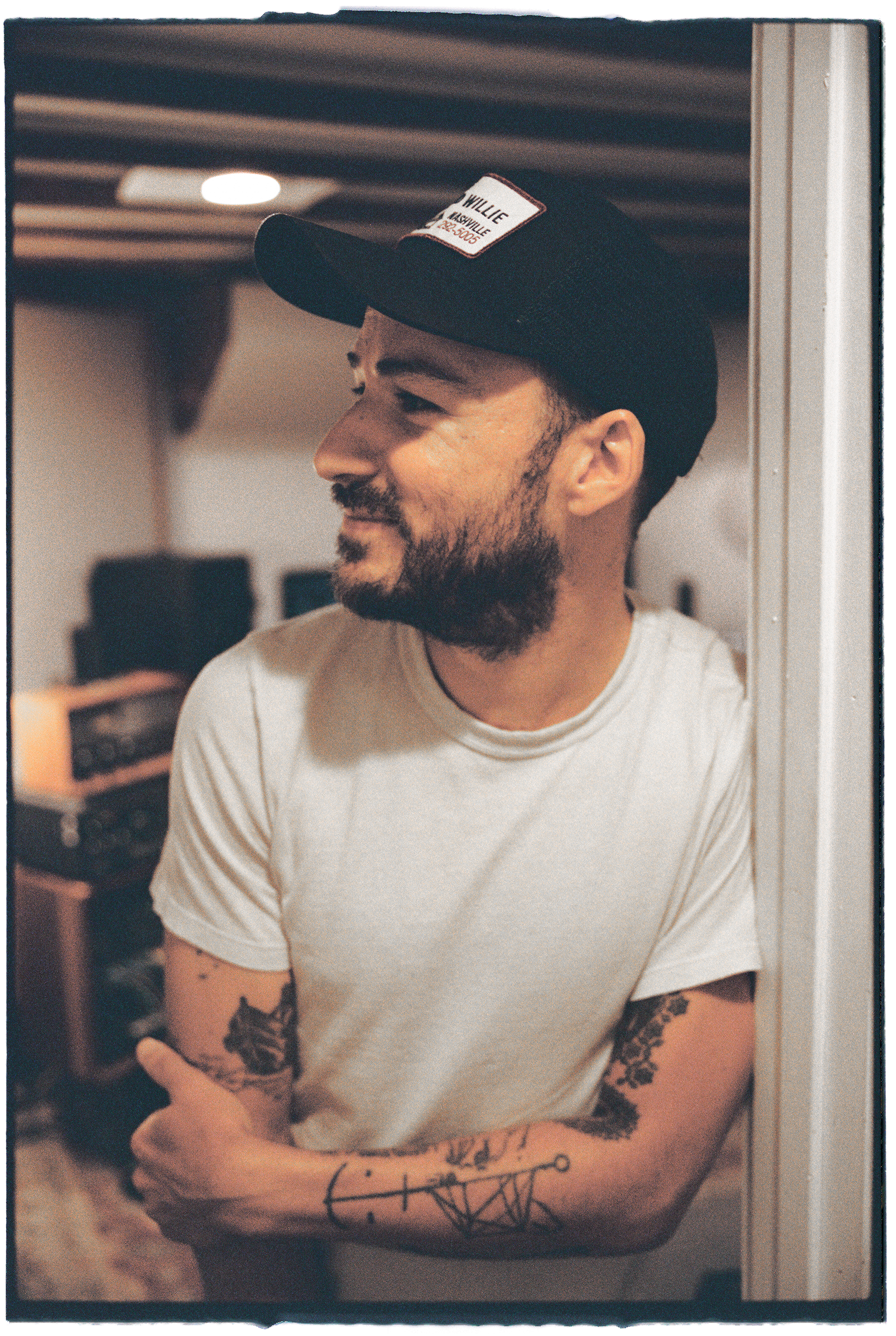 A man with a beard and tattoos on his arms is smiling and looking to his left, standing indoors with his arms crossed, wearing a white t-shirt and a black cap.