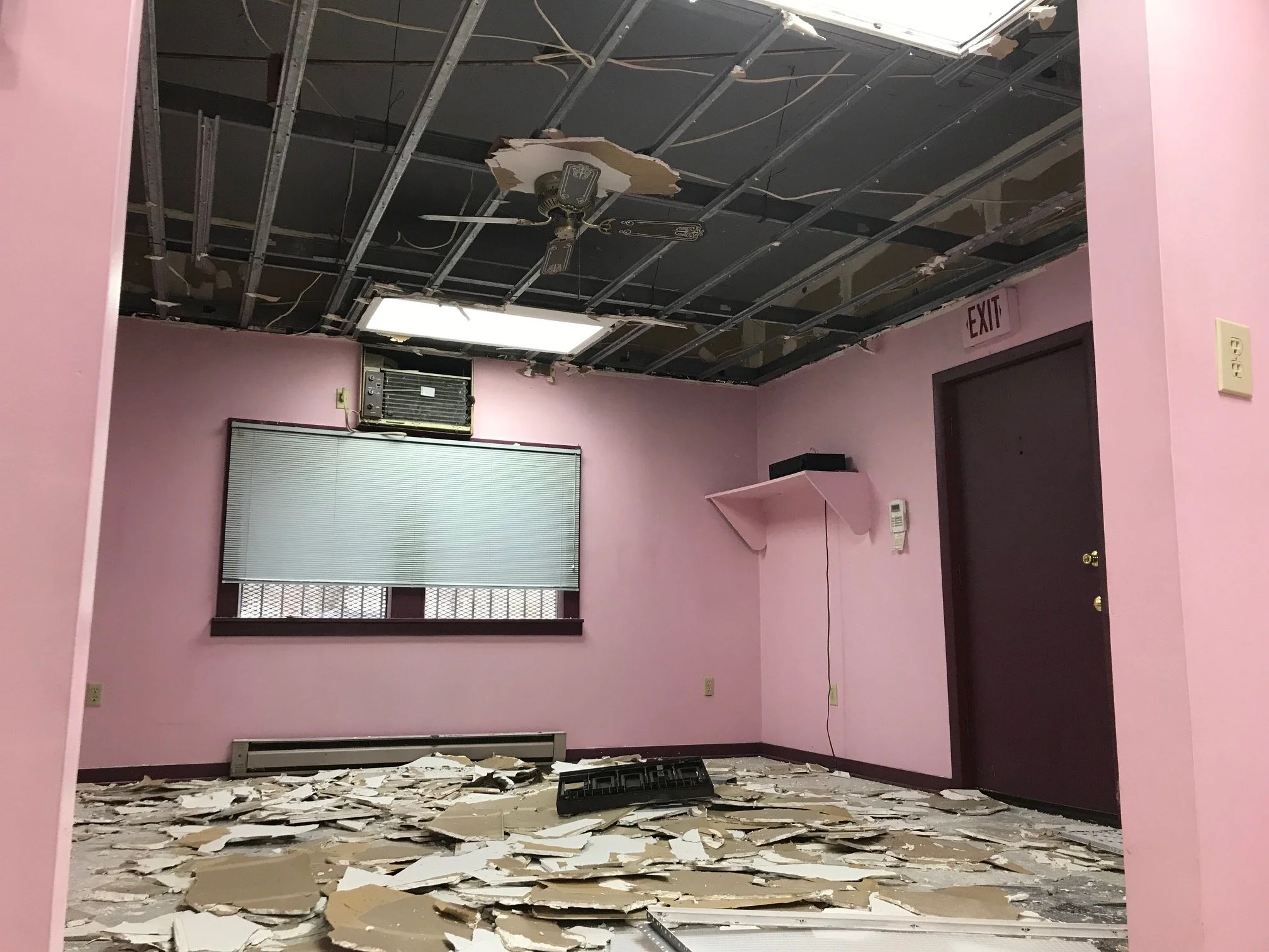 An abandoned room with pink walls and a damaged ceiling, debris and broken plaster on the floor, a window with blinds, an 'EXIT' sign above a door, and an old air conditioning unit.
