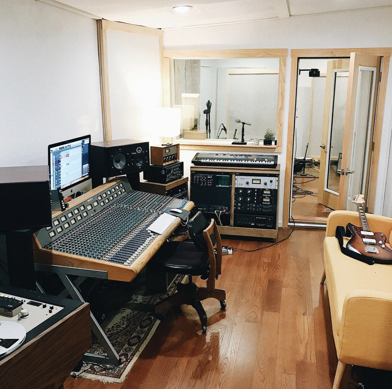 Recording studio with mixing console, computer, speakers, and musical instruments including a guitar, set in a room with wooden walls and flooring.