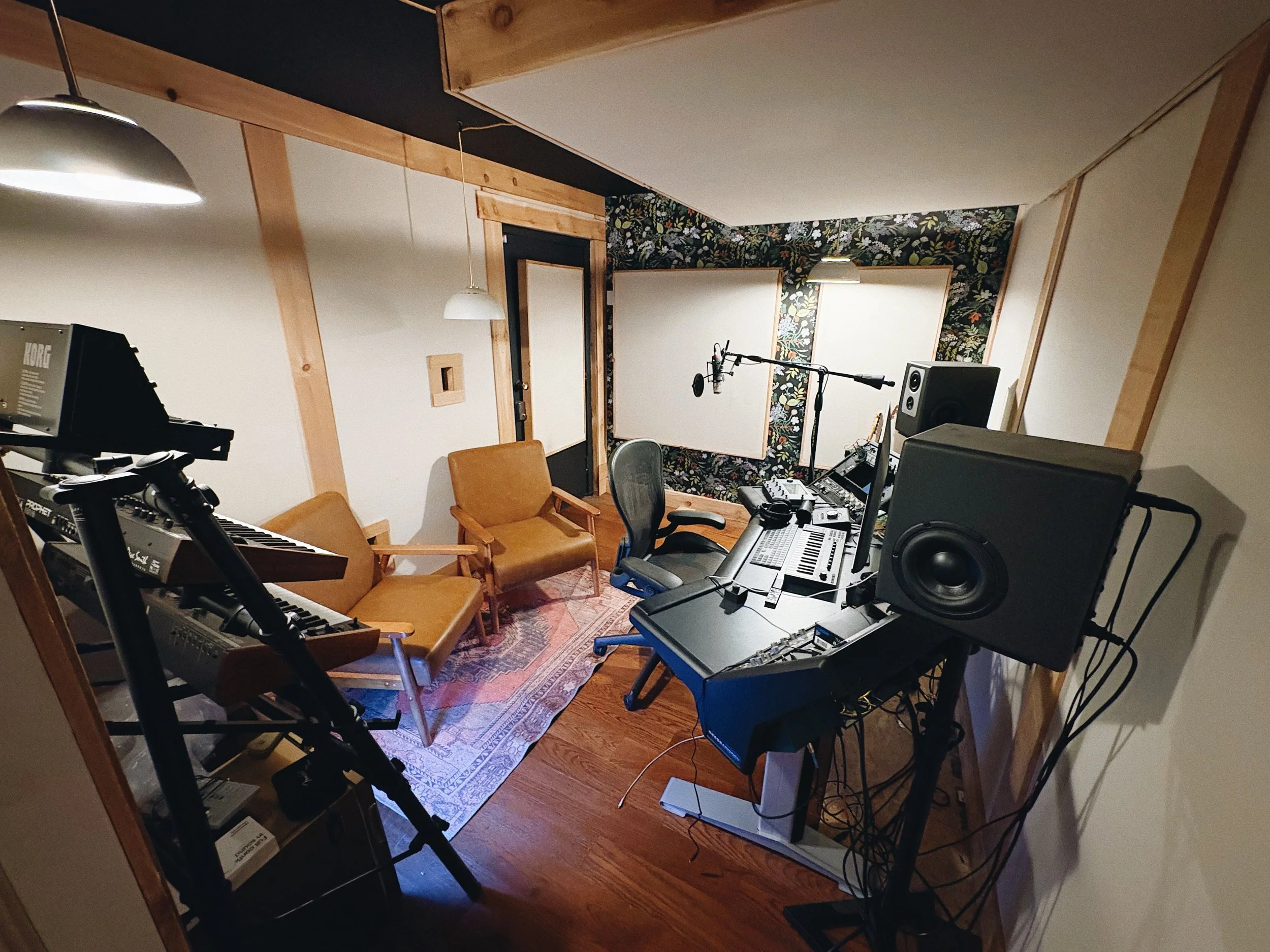 Recording studio with musical and audio production equipment including a keyboard, two chairs, a sound mixer, speaker monitors, a microphone on a stand, and acoustic panels on the wall.