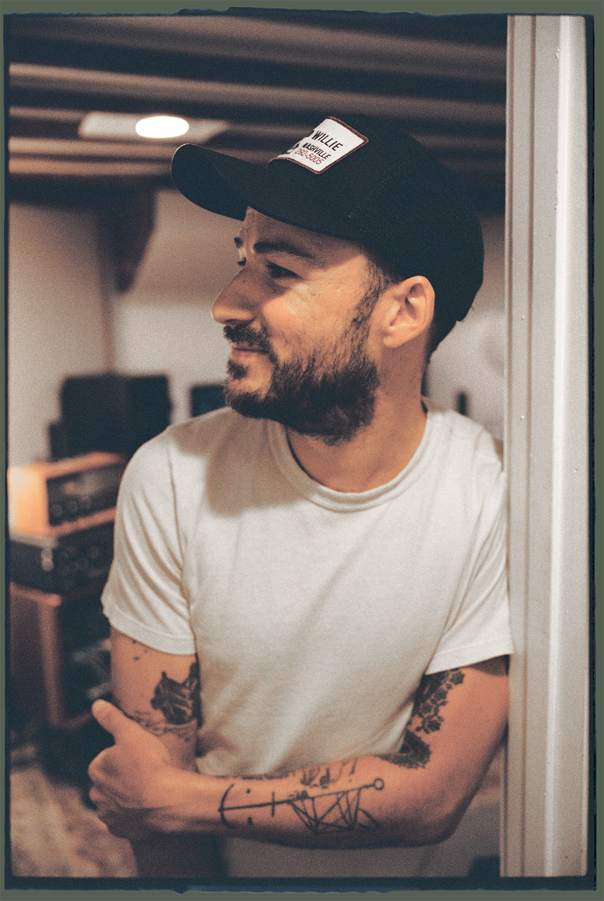 A young man with a beard, wearing a black cap and white t-shirt, standing against a door frame in a room with audio equipment in the background.