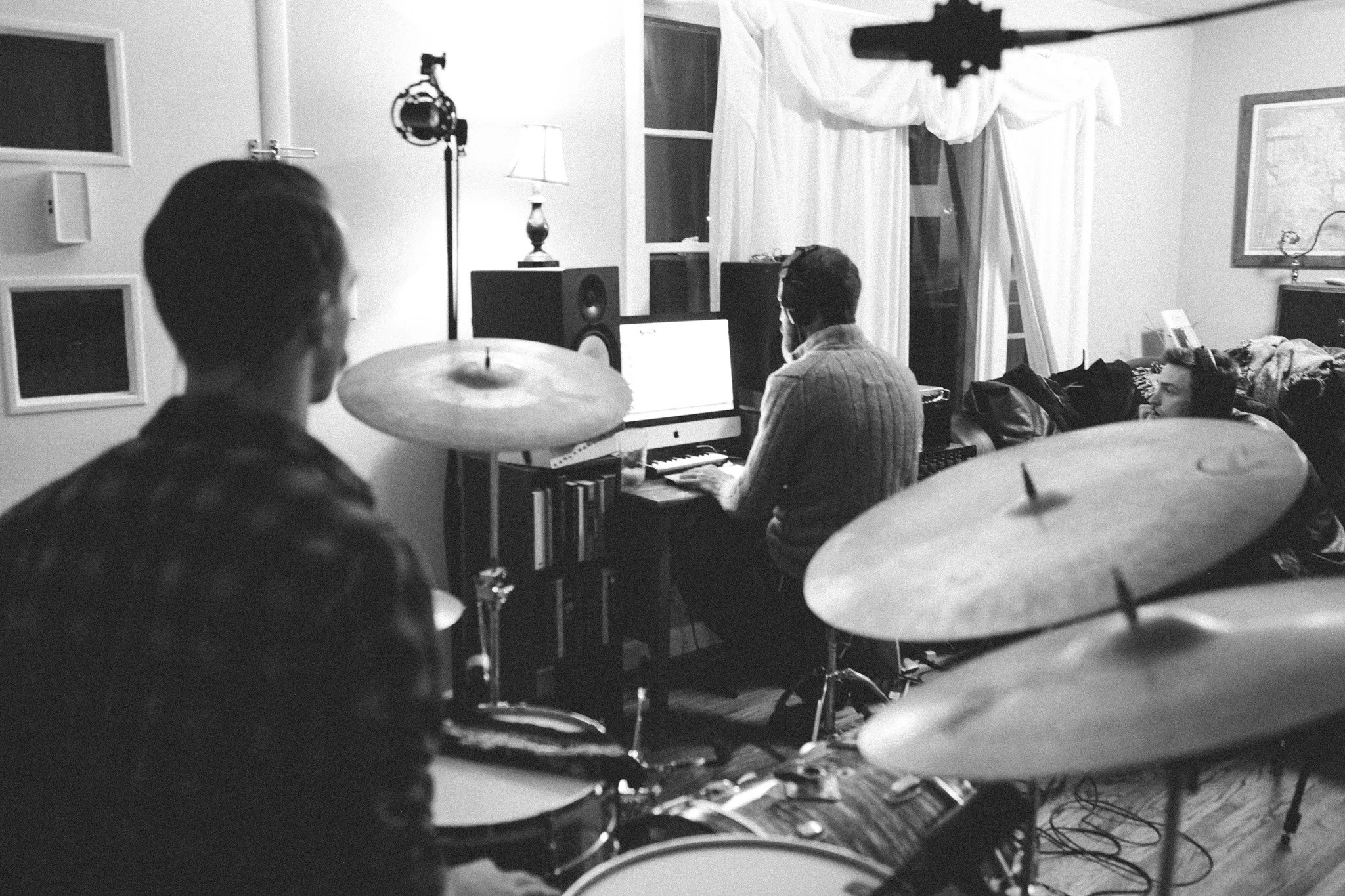 Black and white photo of three people in a home recording studio. One person is playing drums in the foreground, two others are sitting at a desk with music equipment, one on a computer and the other on a keyboard. Studio monitors, soundproof windows, a lamp, and a camera mounted on a microphone stand are visible.