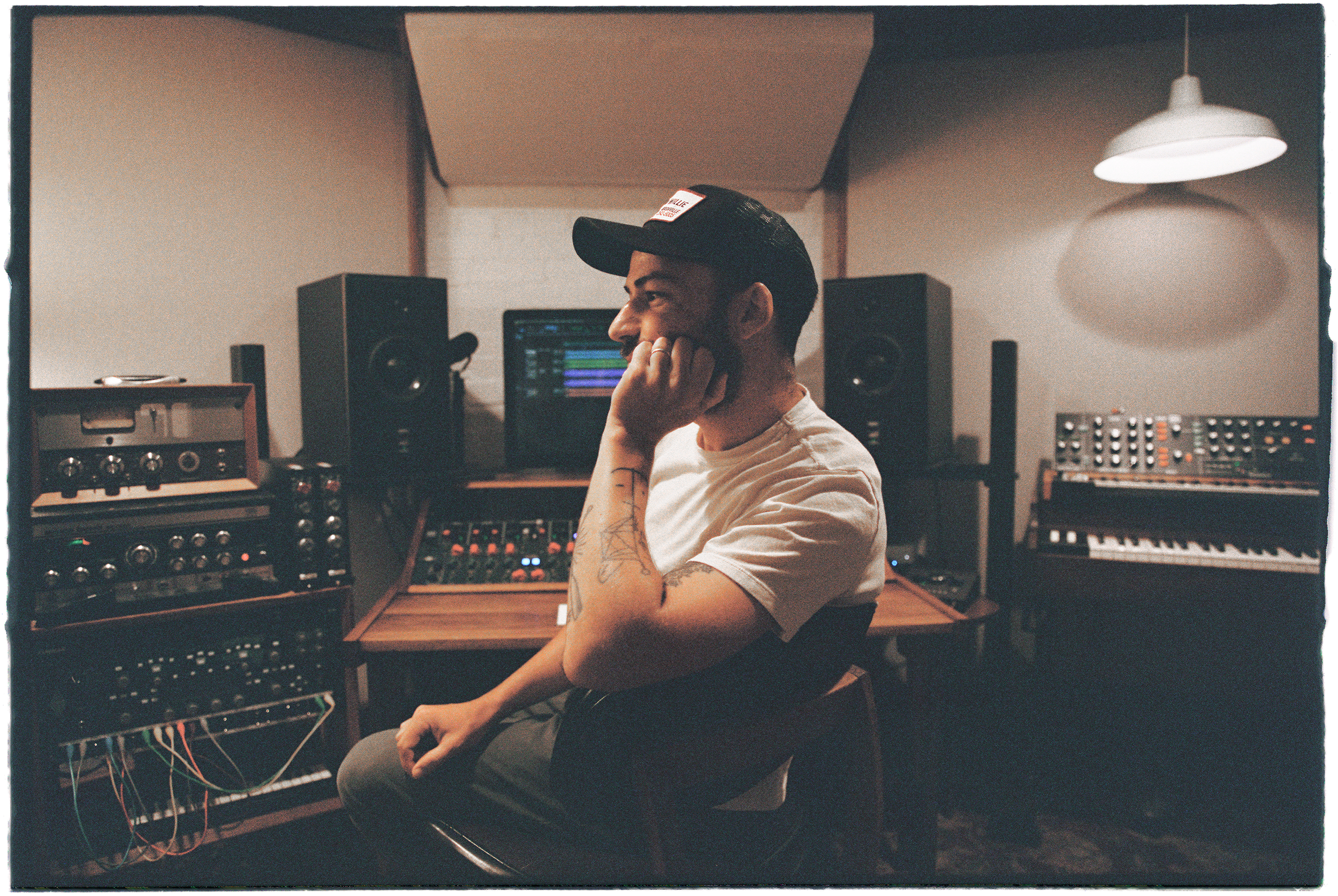 A man sitting in a music studio, wearing a baseball cap and a white t-shirt, surrounded by music equipment including speakers, keyboards, and audio gear.
